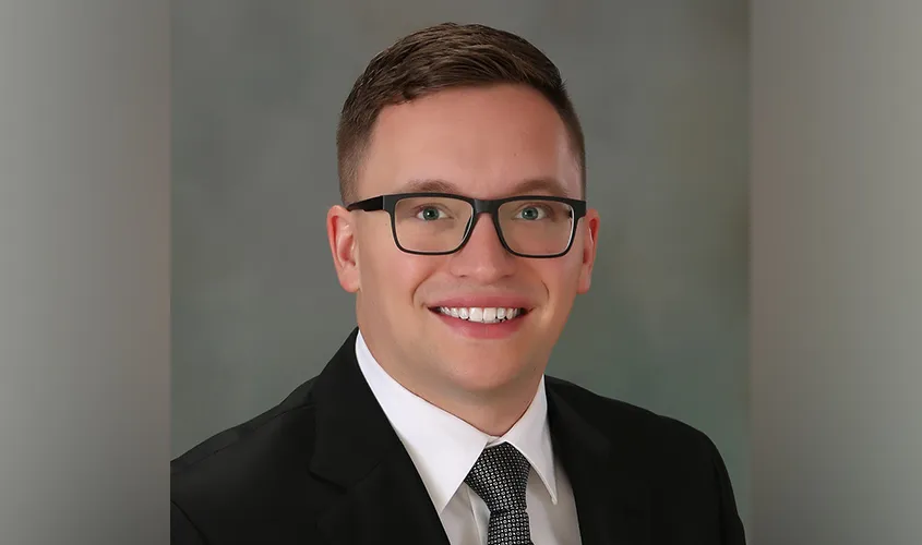 Smiling man with black glasses wearing a suit, white shirt, and tie against a studio background