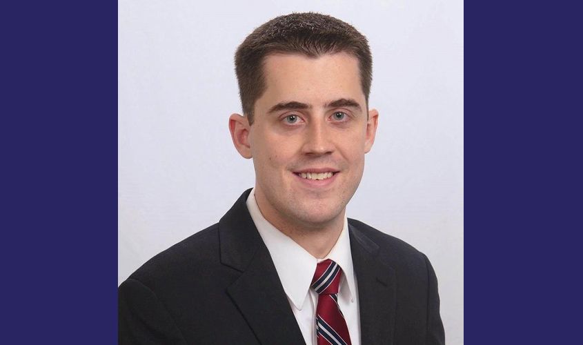 Smiling man with short brown hair wearing a black suit jacket, white collared shirt, and striped red tie News