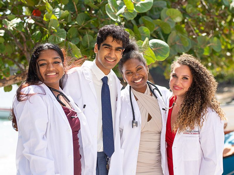 Four smiling medical students wearing white lab coats and stethoscopes outdoors