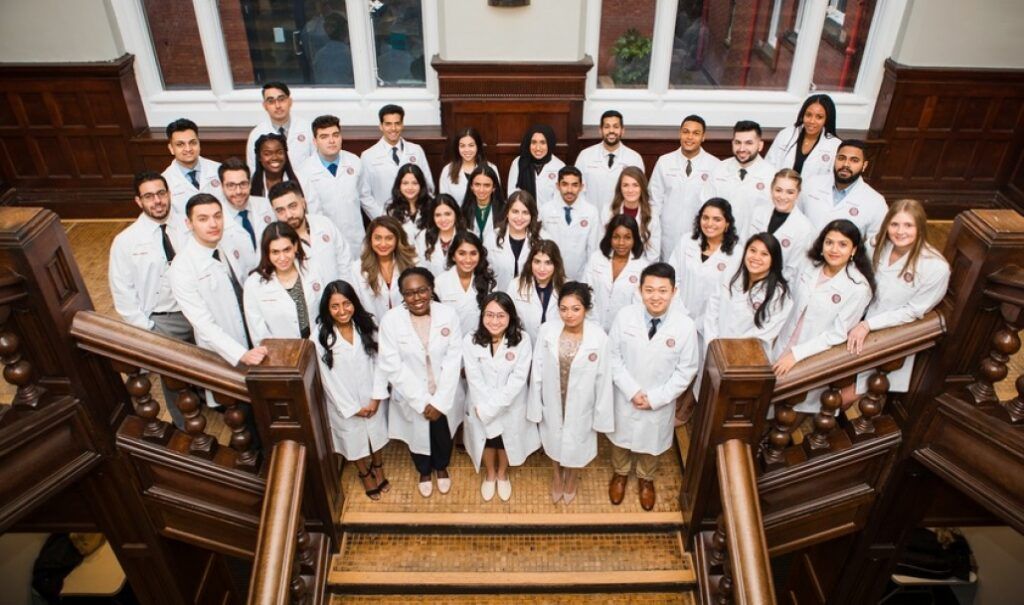 A diverse group of medical students wearing white lab coats poses together on an ornate wooden staircase