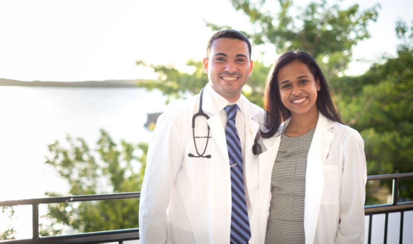 Two St. George's University (SGU) medical students or physicians in white coats and stethoscopes, smiling outdoors.