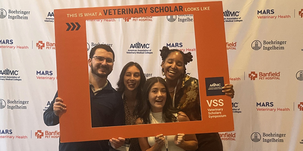 Four attendees pose with an orange photo frame reading "This is what a veterinary scholar looks like" at a symposium