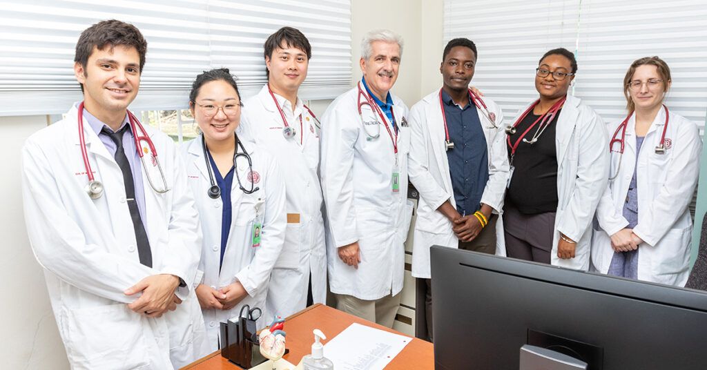 Diverse medical students and a doctor in white lab coats and stethoscopes posing together in a clinic office
