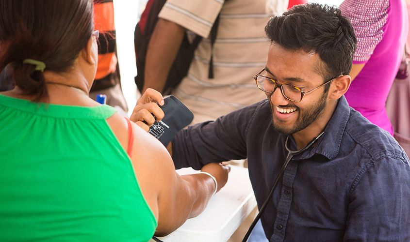 Medical student wearing a stethoscope smiles as he measures a woman's blood pressure during a community health clinic screening.