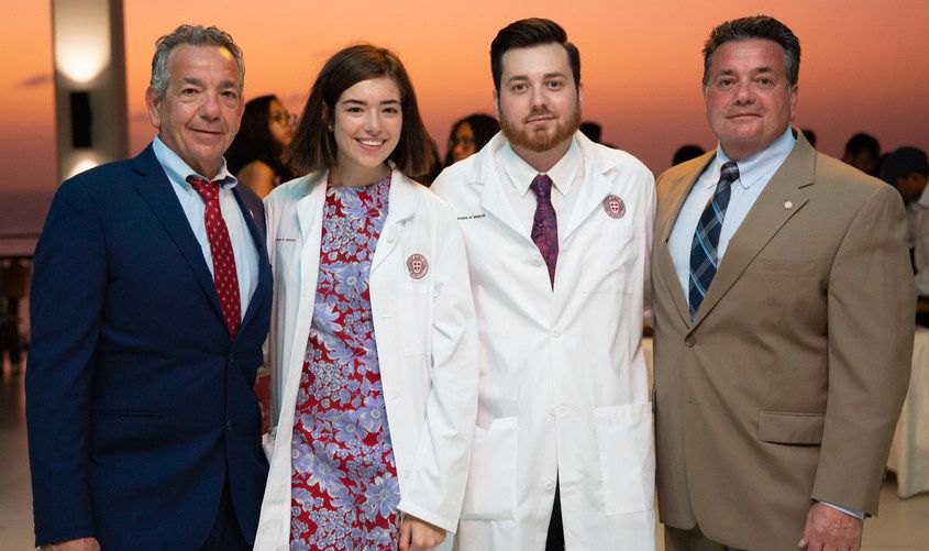 Two medical students wearing white coats stand between two men in suits at a sunset reception News