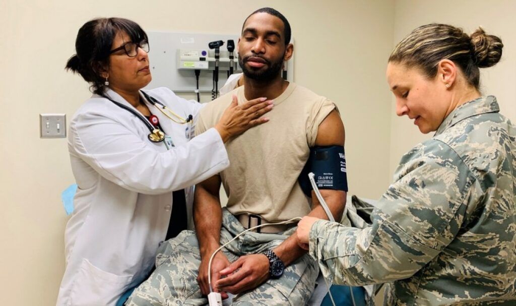 A doctor checks a servicemember's neck while a military medic monitors his vitals in an exam room