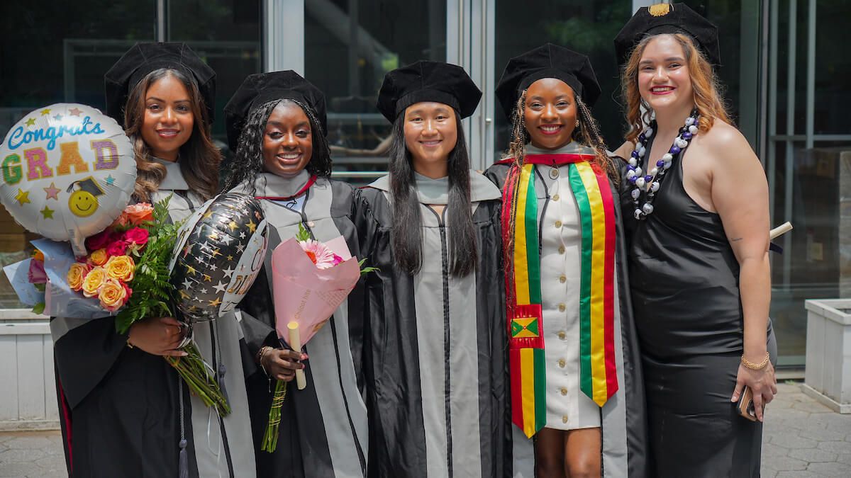 Five smiling graduates in academic regalia holding bouquets of flowers and celebratory balloons