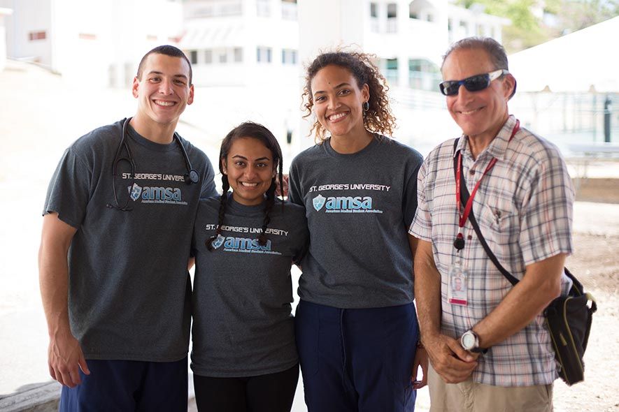 Three St. George's University medical students in AMSA shirts smile outdoors alongside a colleague