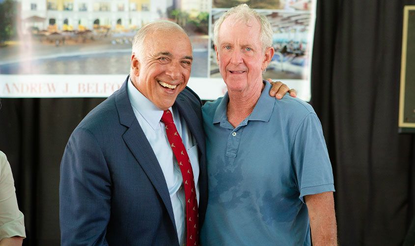 Two smiling men pose together indoors, one in a suit with a red tie and the other in a blue polo shirt