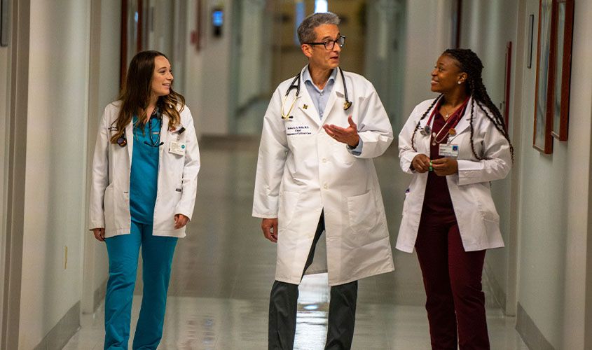 Three doctors in lab coats and scrubs talking while walking down a hospital hallway
