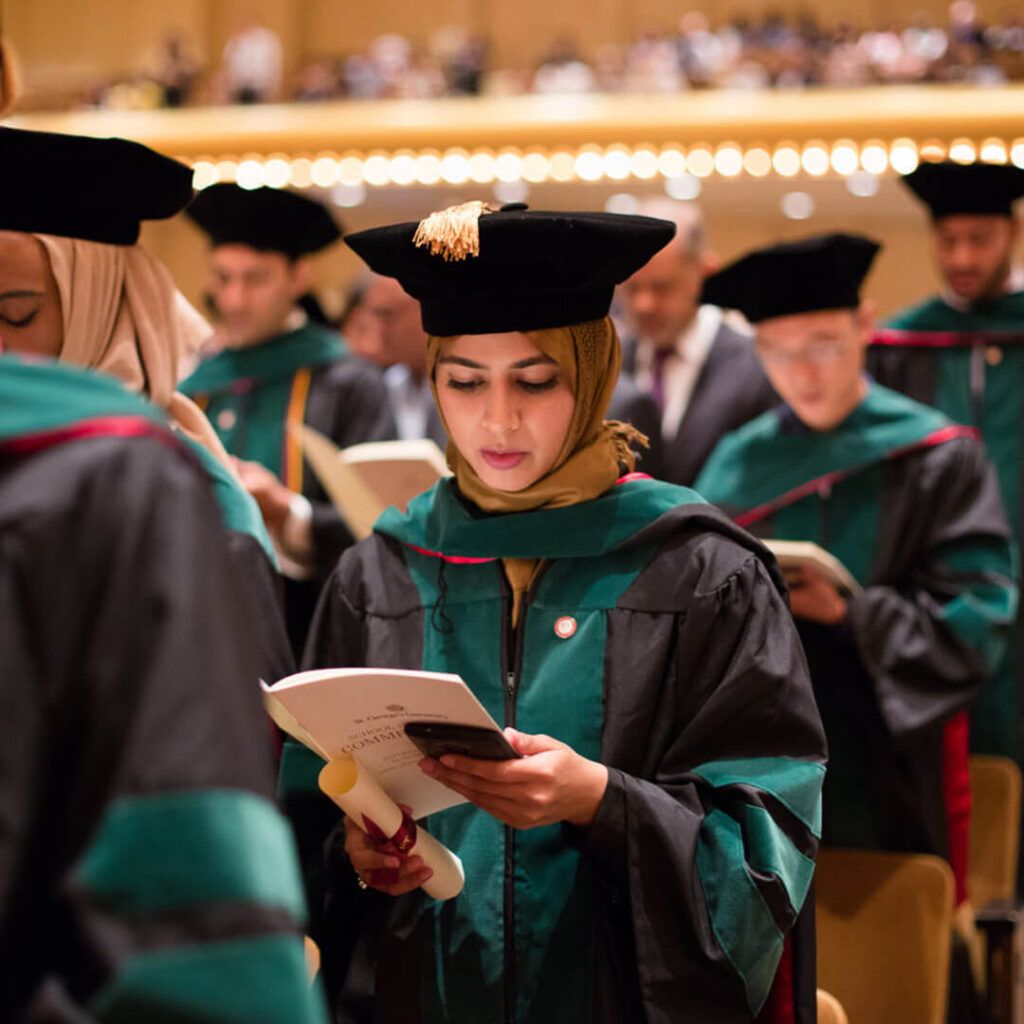 A graduate in a hijab and academic regalia looks at her phone while holding a commencement program