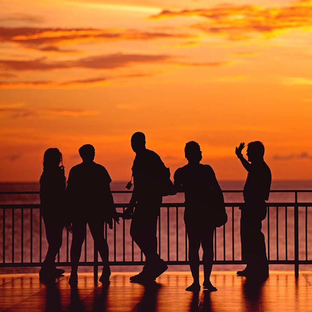 Silhouettes of five people standing by a railing overlooking the ocean against a vibrant orange sunset sky
