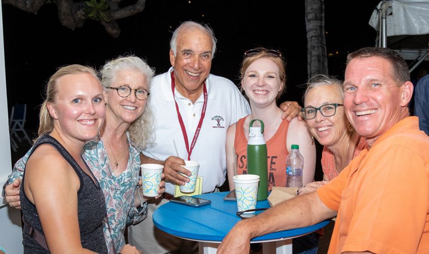 Six smiling people pose together around an outdoor table at an evening social gathering