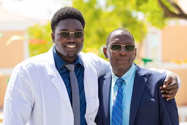 A smiling man in a white lab coat with his arm around a man wearing a suit outdoors