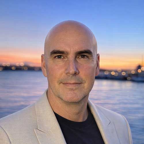 Bald man in a light-colored blazer standing in front of a waterfront at sunset