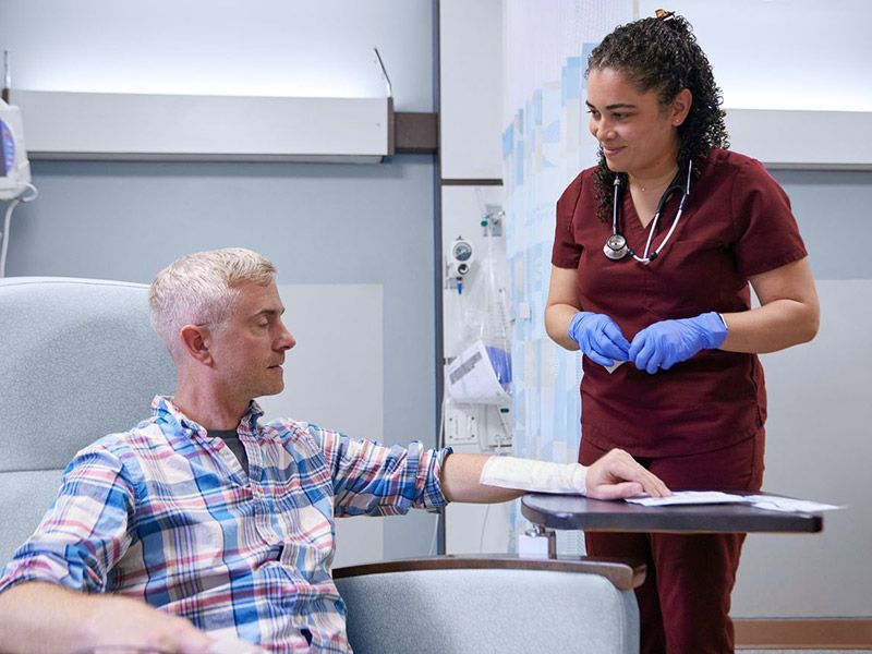 A nurse in maroon scrubs attends to a seated patient with a bandaged arm