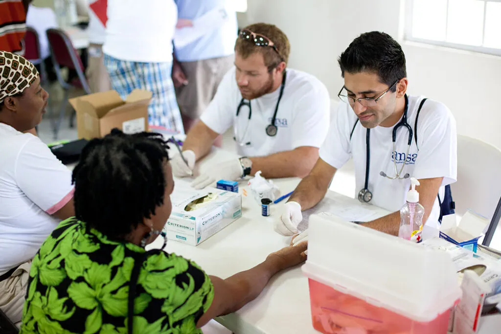 Two medical volunteers with stethoscopes perform health screenings for patients seated across a table