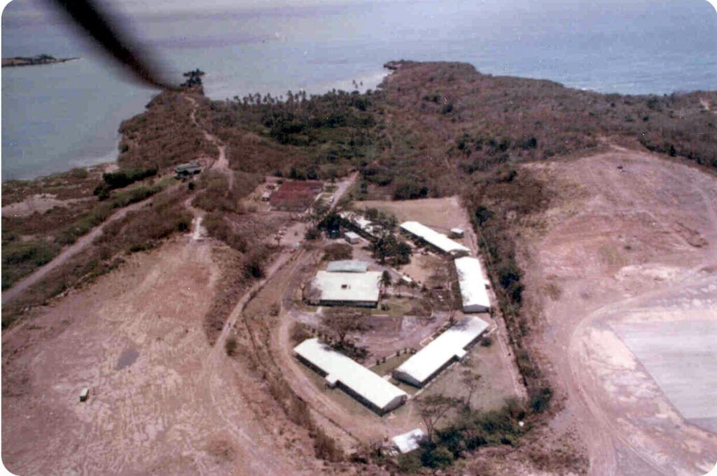 Aerial view of a coastal compound with several white-roofed buildings surrounded by cleared land next to the sea