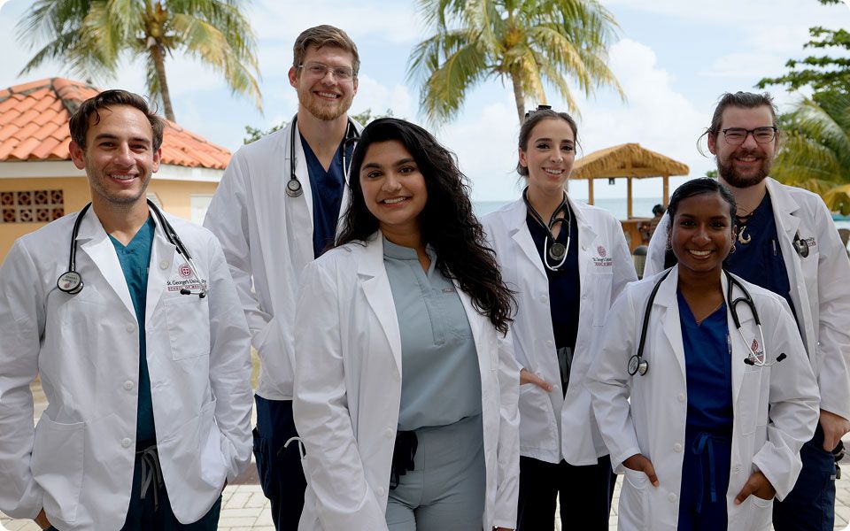 Six smiling St. George's University medical students wearing white lab coats and stethoscopes outdoors