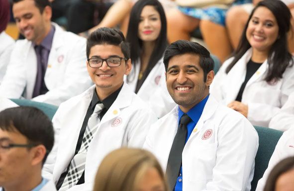 Medical students wearing white coats and ties smiling in an auditorium