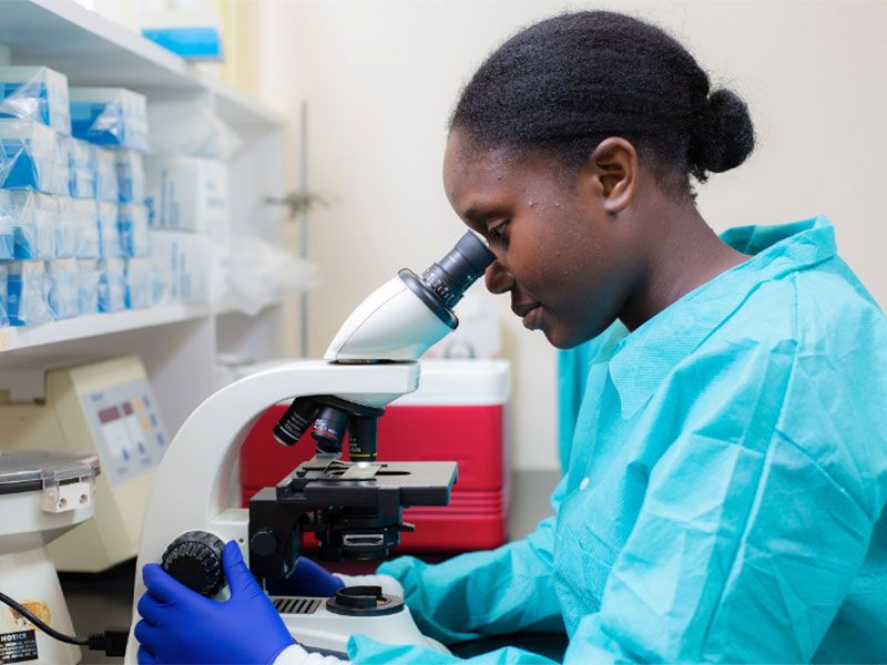 A lab researcher wearing protective scrubs and gloves looks closely into a microscope