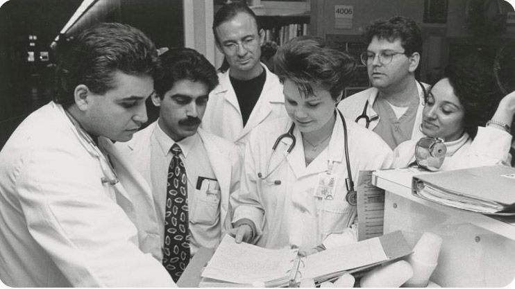 Doctors in white lab coats gather around a hospital desk to review notes in an open binder