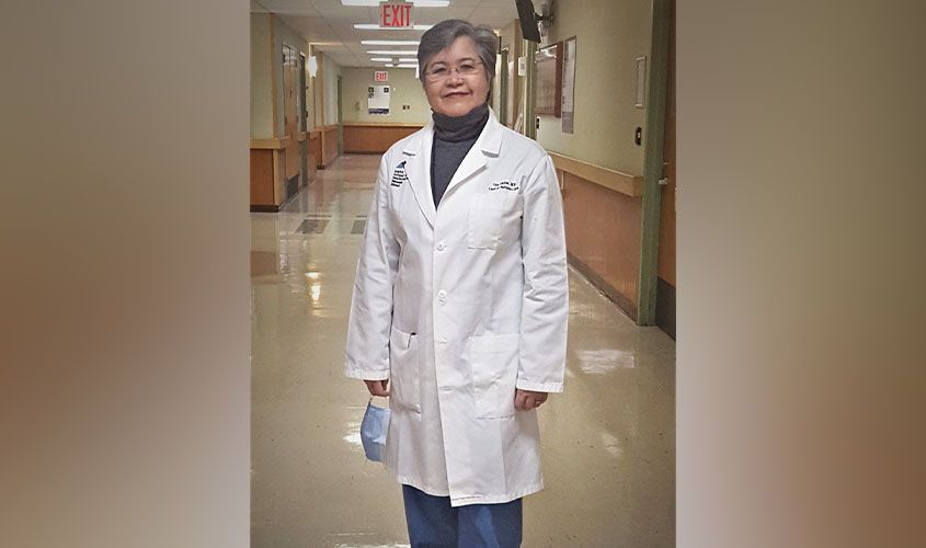 Doctor wearing glasses and a white lab coat standing in a hospital hallway holding a face mask