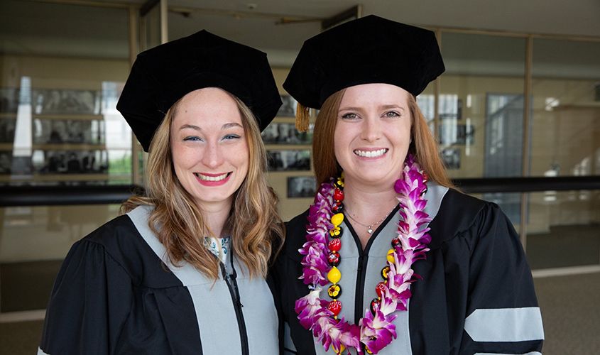 Two SGU female graduates smiling in doctoral tams and academic gowns, one wearing a lei, at their commencement ceremony.