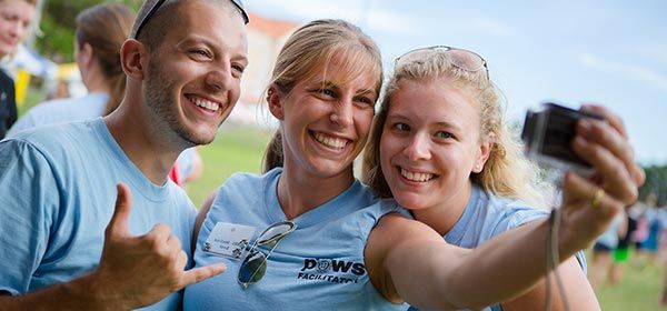 Three smiling event facilitators in light blue shirts pose together for a selfie outdoors