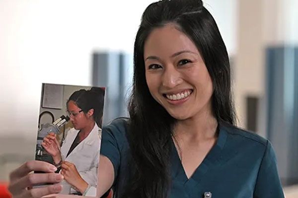 A smiling woman in scrubs holds up a photo of herself wearing a lab coat and using a microscope